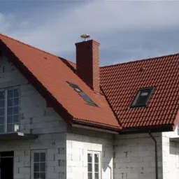 Dom w trakcie budowy z czerwoną dachówką ceramiczną, widoczne okna dachowe i komin z anteną, ściany z białego bloczka.